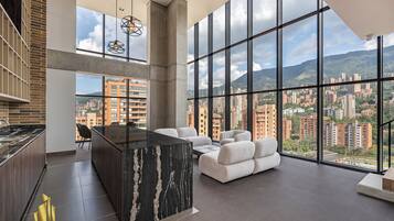 Una cocina moderna con una isla de mármol, un área de comedor con sofás blancos y ventanas amplias con vistas a la ciudad.