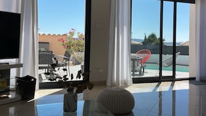 Un salon spacieux doté d’un grand téléviseur, d’une table basse en verre et d’un coin salon confortable avec un fauteuil rouge et un pouf blanc.