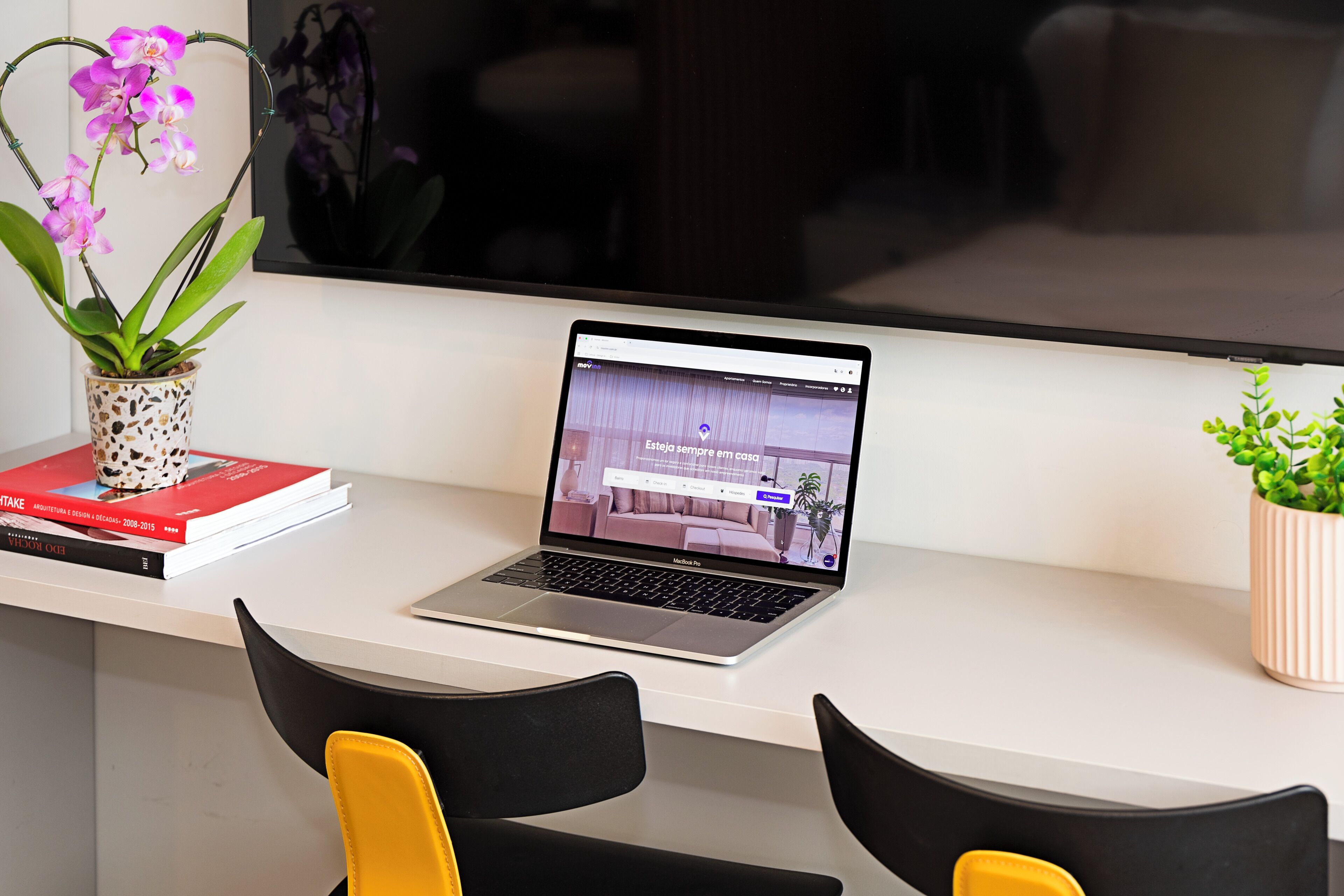 A laptop on a desk with a potted orchid, a book, and a small plant.