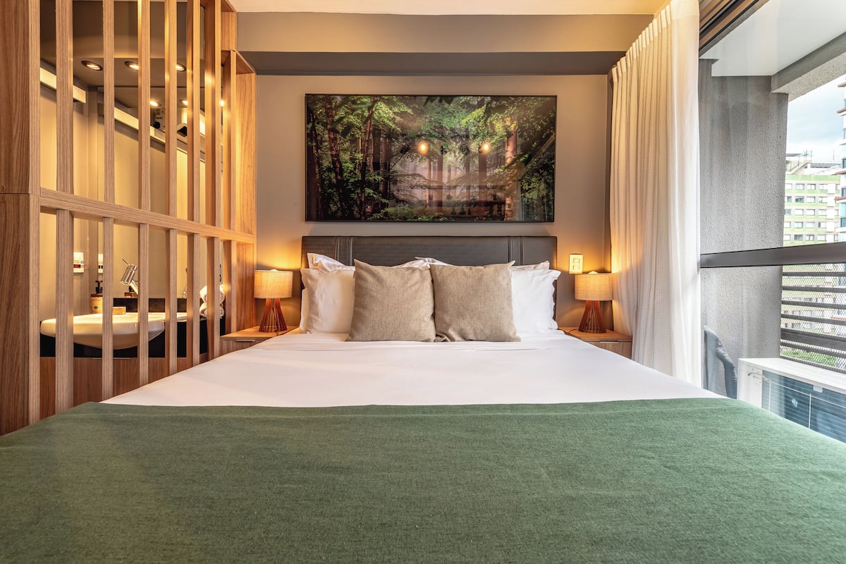 A hotel room with a large bed, a painting of a forest, a view of buildings, and a balcony.