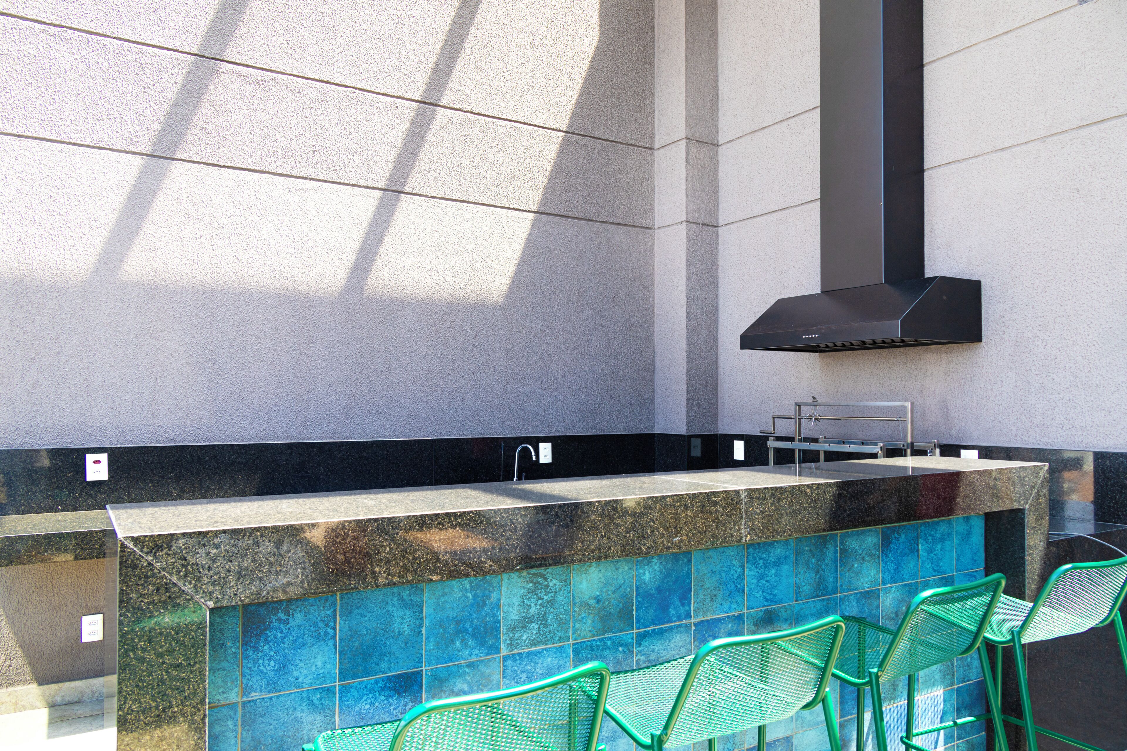 A modern outdoor bar area with a stone countertop, green metal chairs, and a black range hood.
