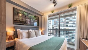 A hotel room with a large bed, a painting above the bed, a nightstand with a lamp, a window with curtains, and a view of a building outside. - Movinn Nine - Jardim Paulista (São Paulo)