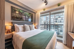 Une chambre d’hôtel avec un grand lit, un tableau au-dessus du lit, une table de chevet avec une lampe, une fenêtre avec des rideaux et une vue sur un bâtiment à l’extérieur.