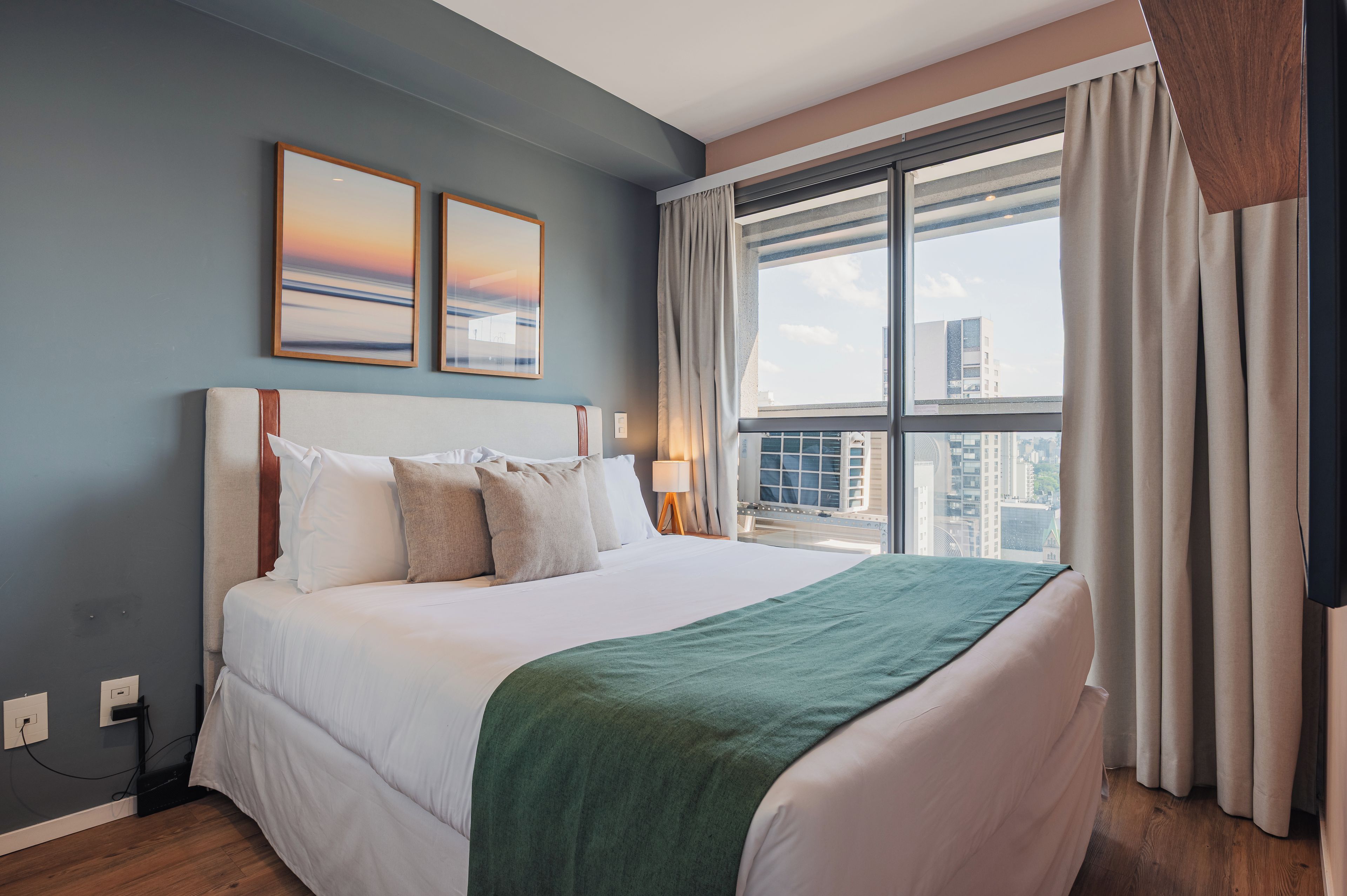 Quarto de hotel com uma cama grande, duas pinturas penduradas na parede, uma janela com cortinas e vista para edifícios.