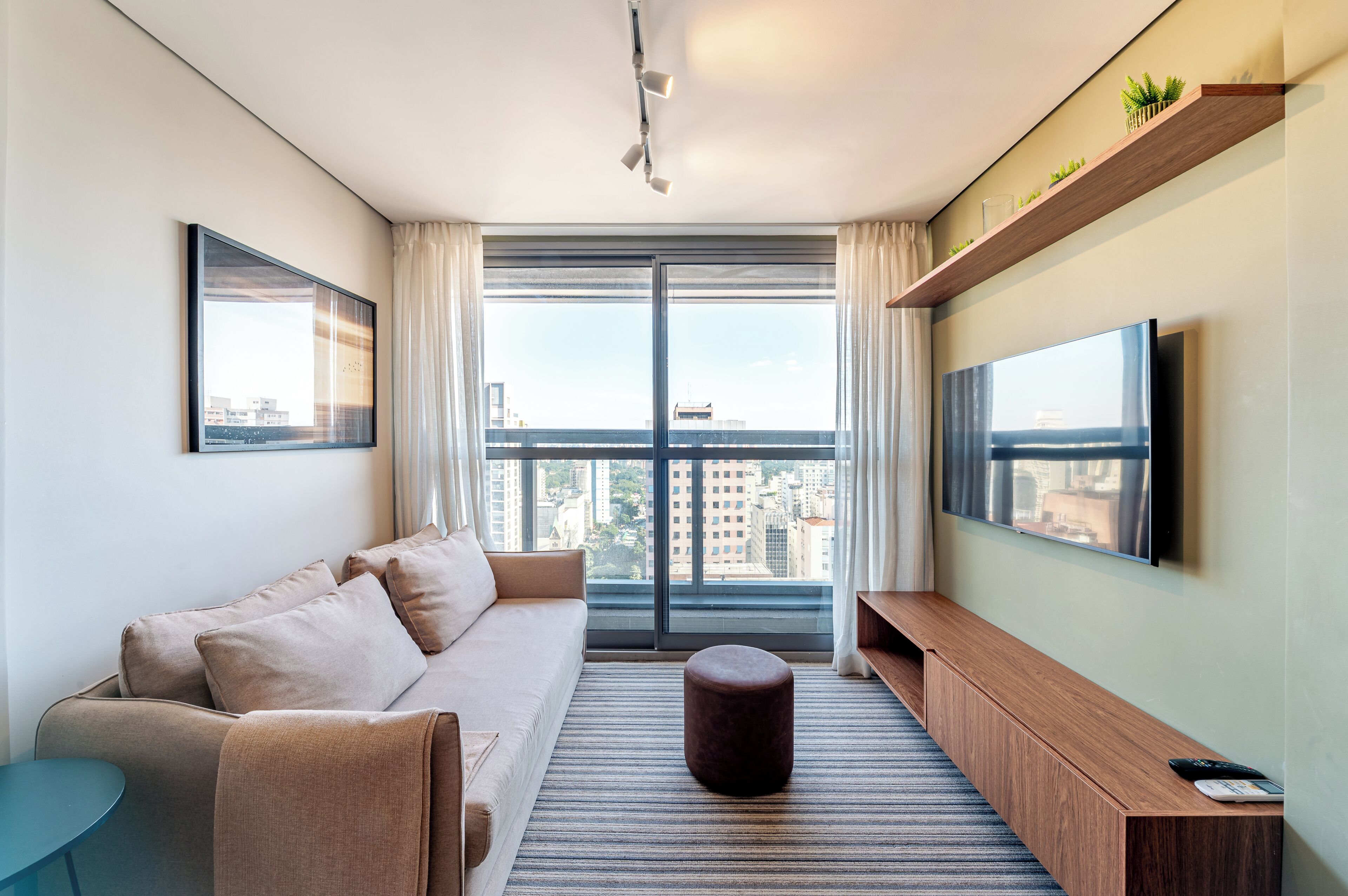 A modern living room with a sofa, ottoman, TV, and a large window with a city view.