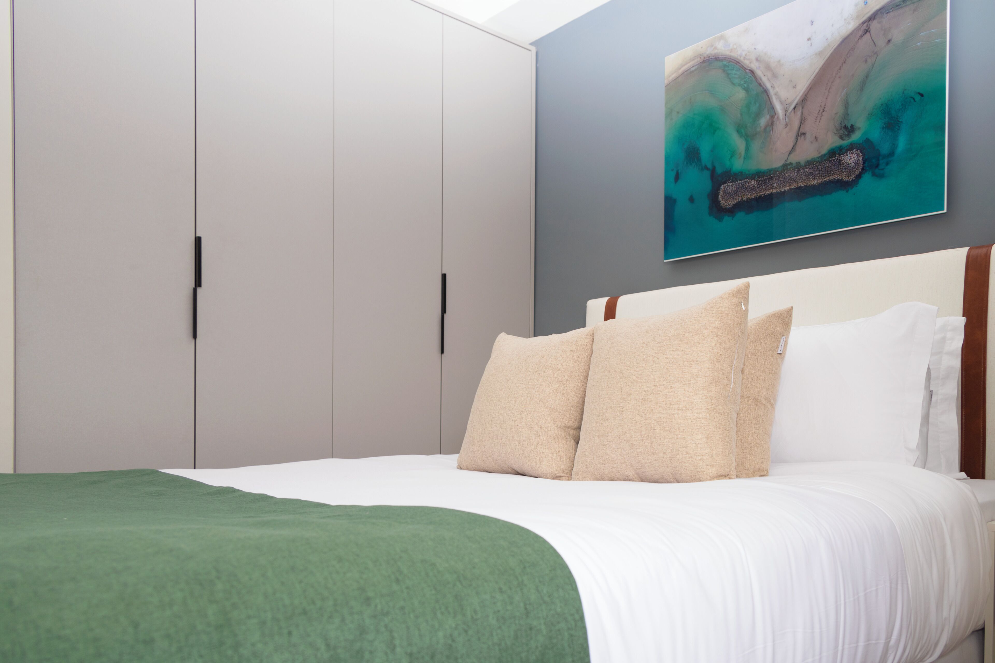 Ein Schlafzimmer mit einem Bett, einer grünen Tagesdecke, beigen Kissen und einem großen abstrakten Gemälde an der Wand.
