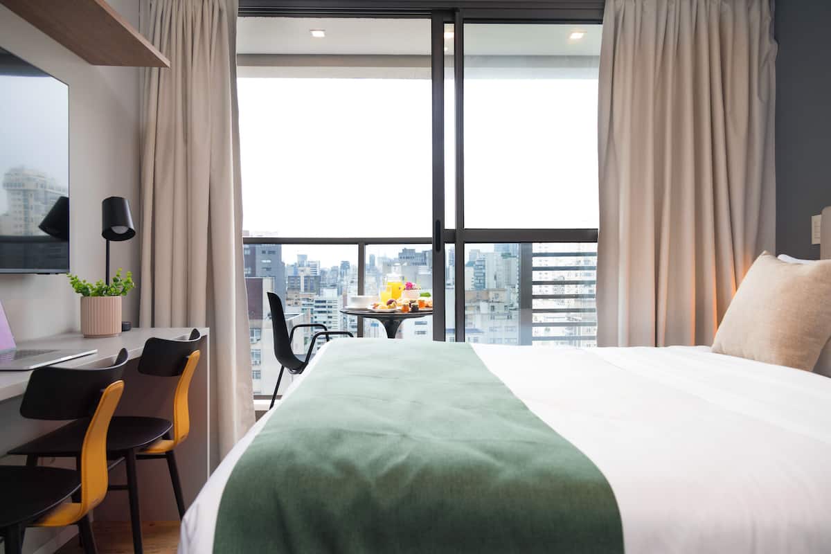 A hotel room with a bed, a desk, chairs, a small table with fruits, and a city view through large windows.
