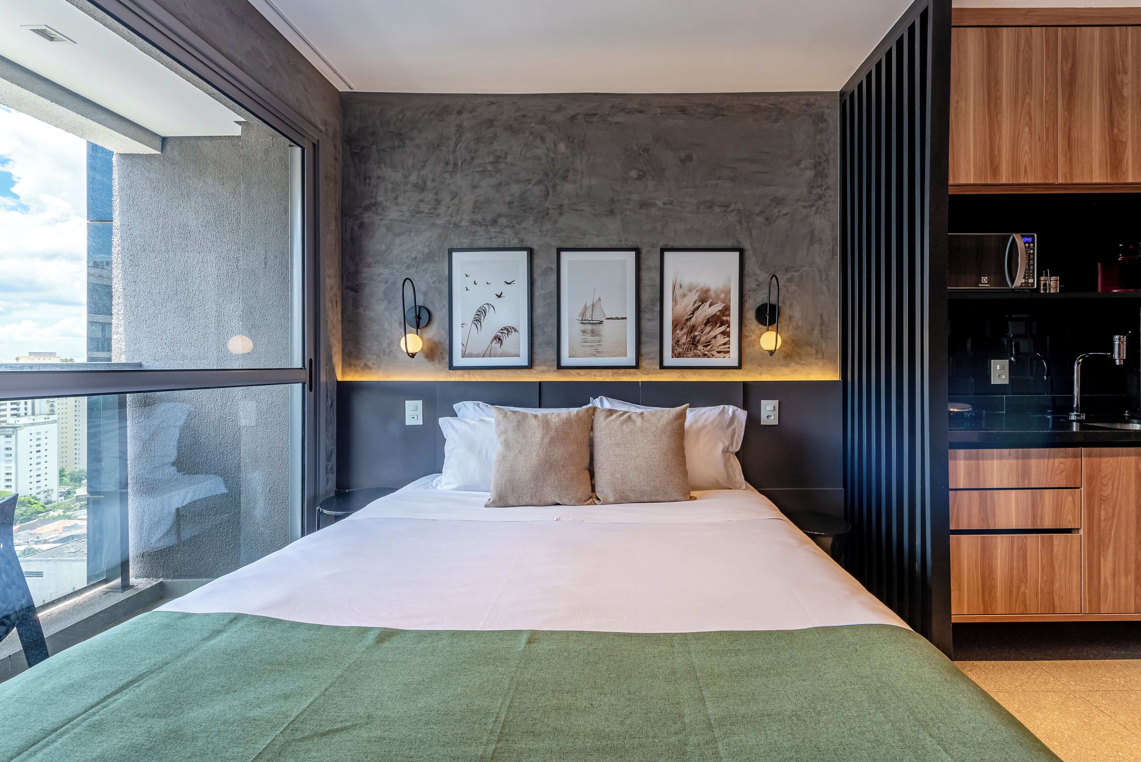 A modern bedroom with a large bed, a view of the city, and a kitchenette.