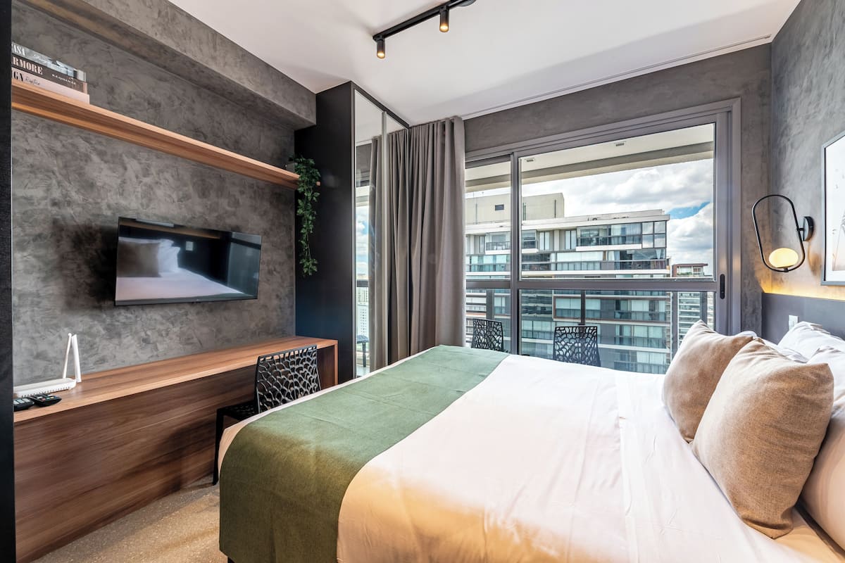 A modern hotel room with a large bed, a flat-screen TV mounted on the wall, a desk with a chair, and a balcony with a view of the city.