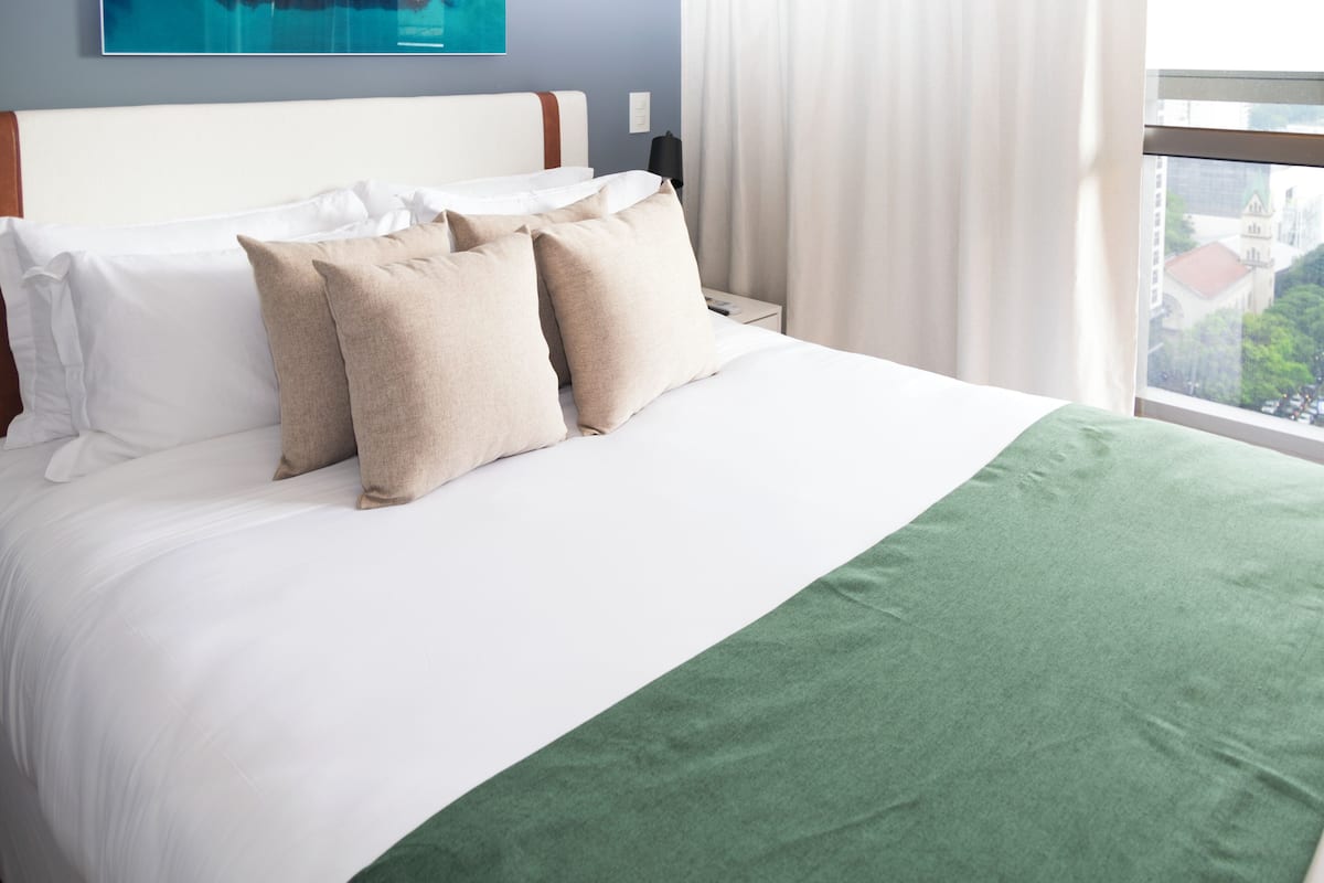 A neatly made bed with white and beige pillows, a green throw blanket, and a view of a cityscape through a window.