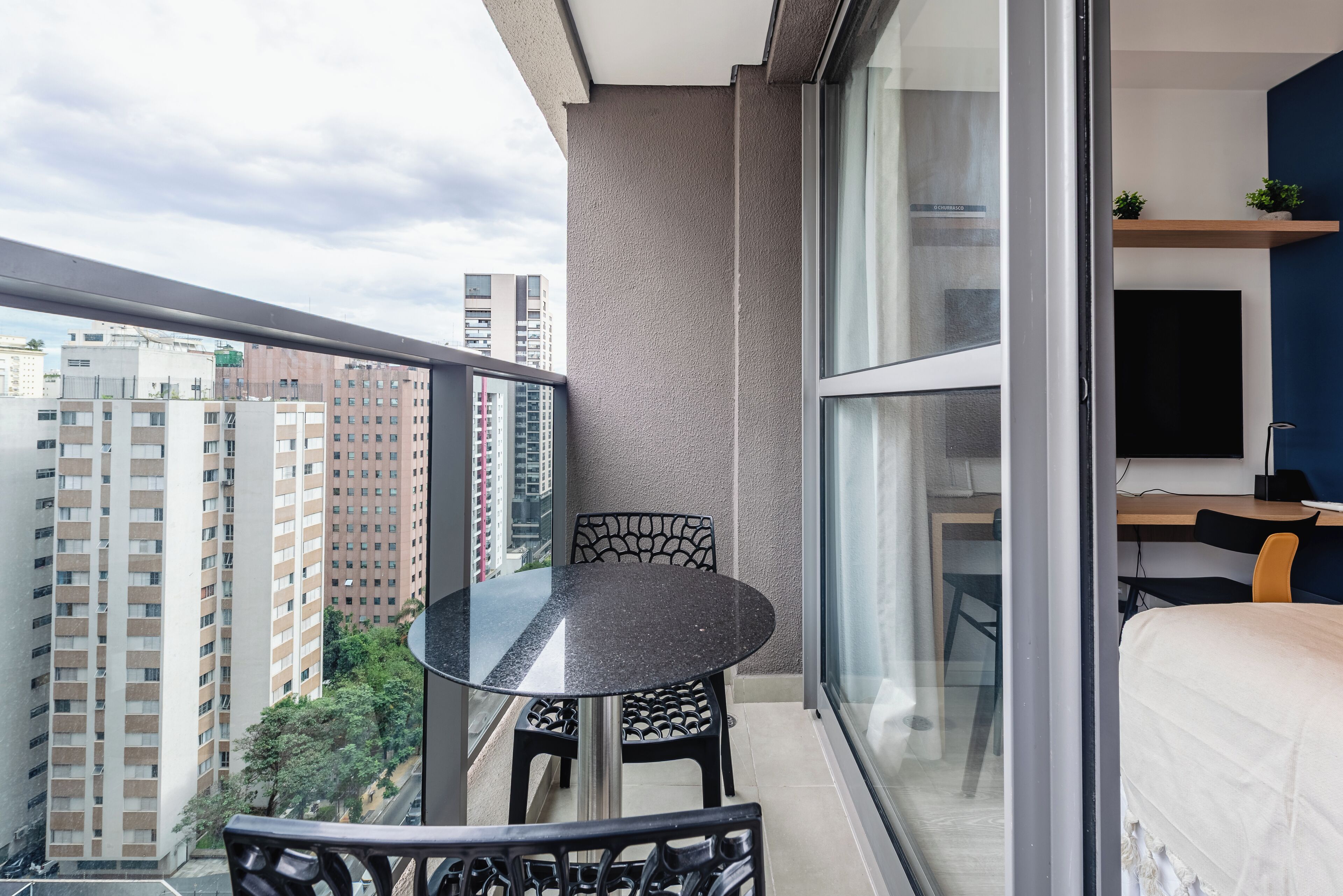Un balcón con una mesa y sillas de vidrio, una cama, un escritorio con una computadora y vistas a edificios altos.