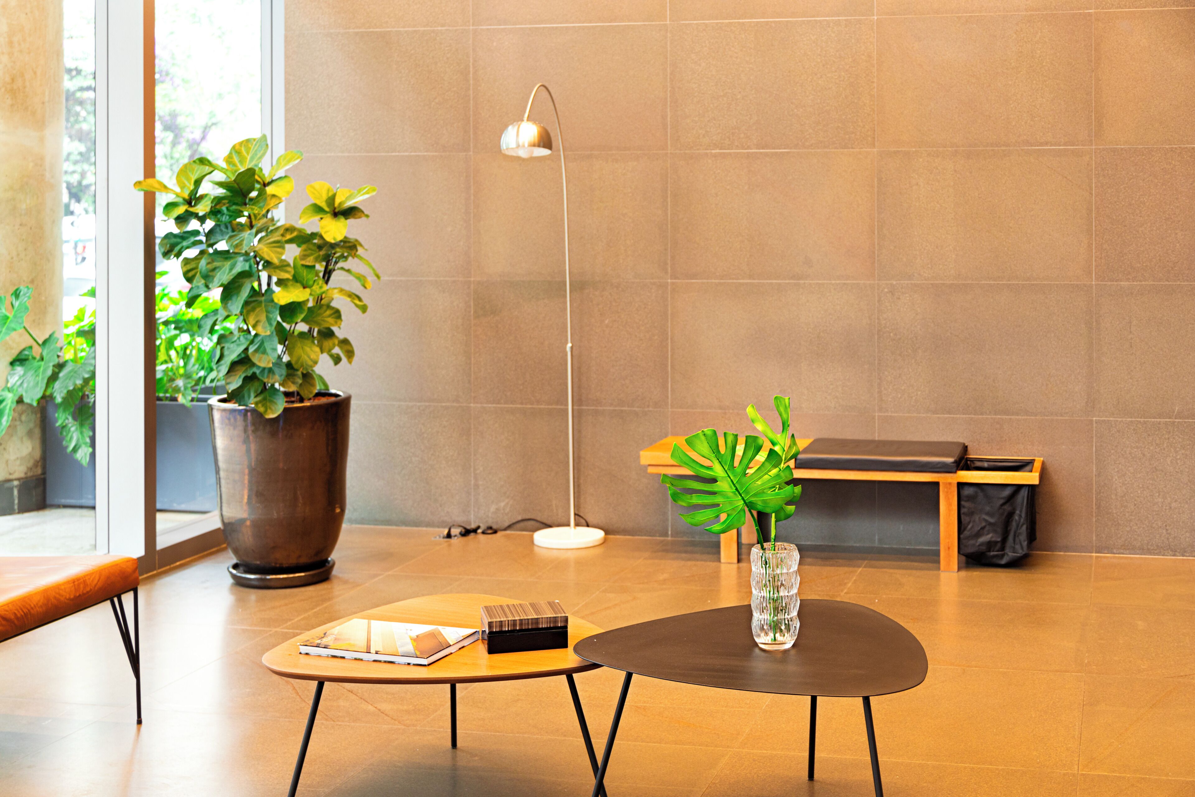A modern lobby with a large potted plant, a round table with a notebook, a floor lamp, and a bench.