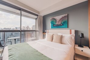 A hotel room with a large bed, a city view, a painting on the wall, and a desk with a lamp.