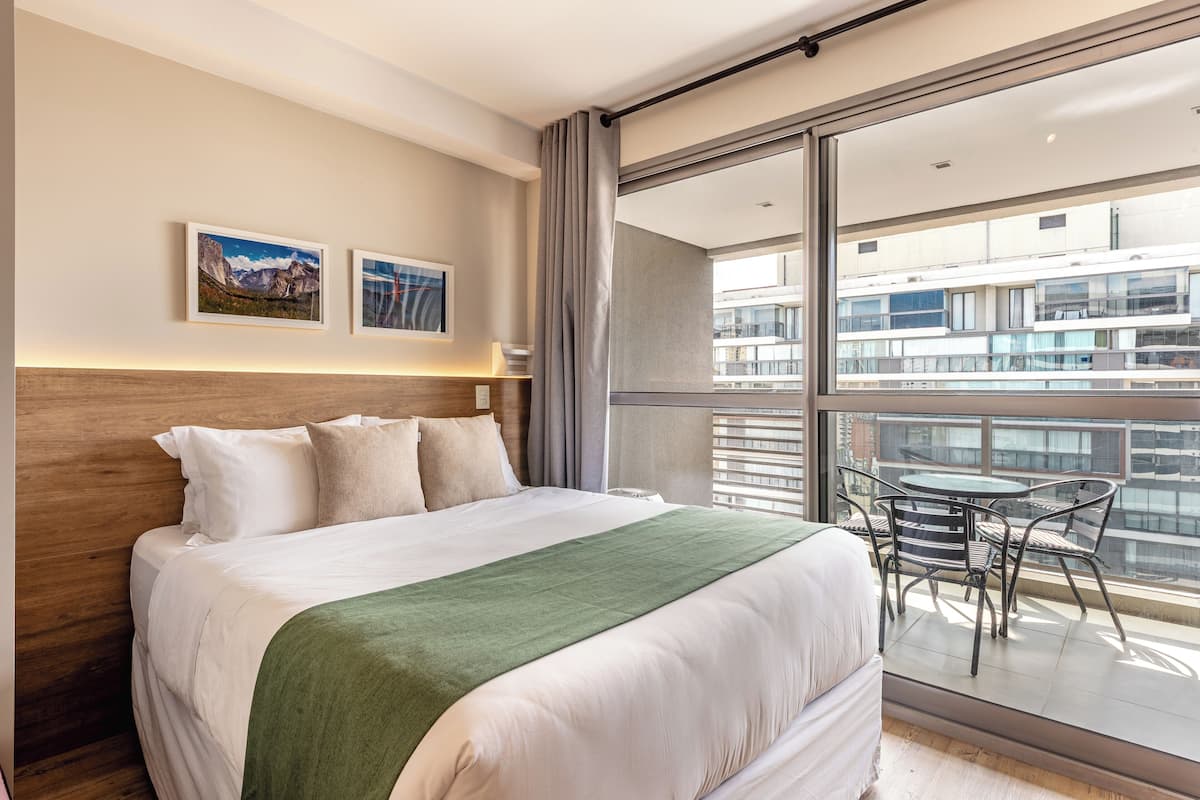 A hotel room with a bed, a green throw, two framed pictures on the wall, a balcony with a table and chairs, and a view of buildings.