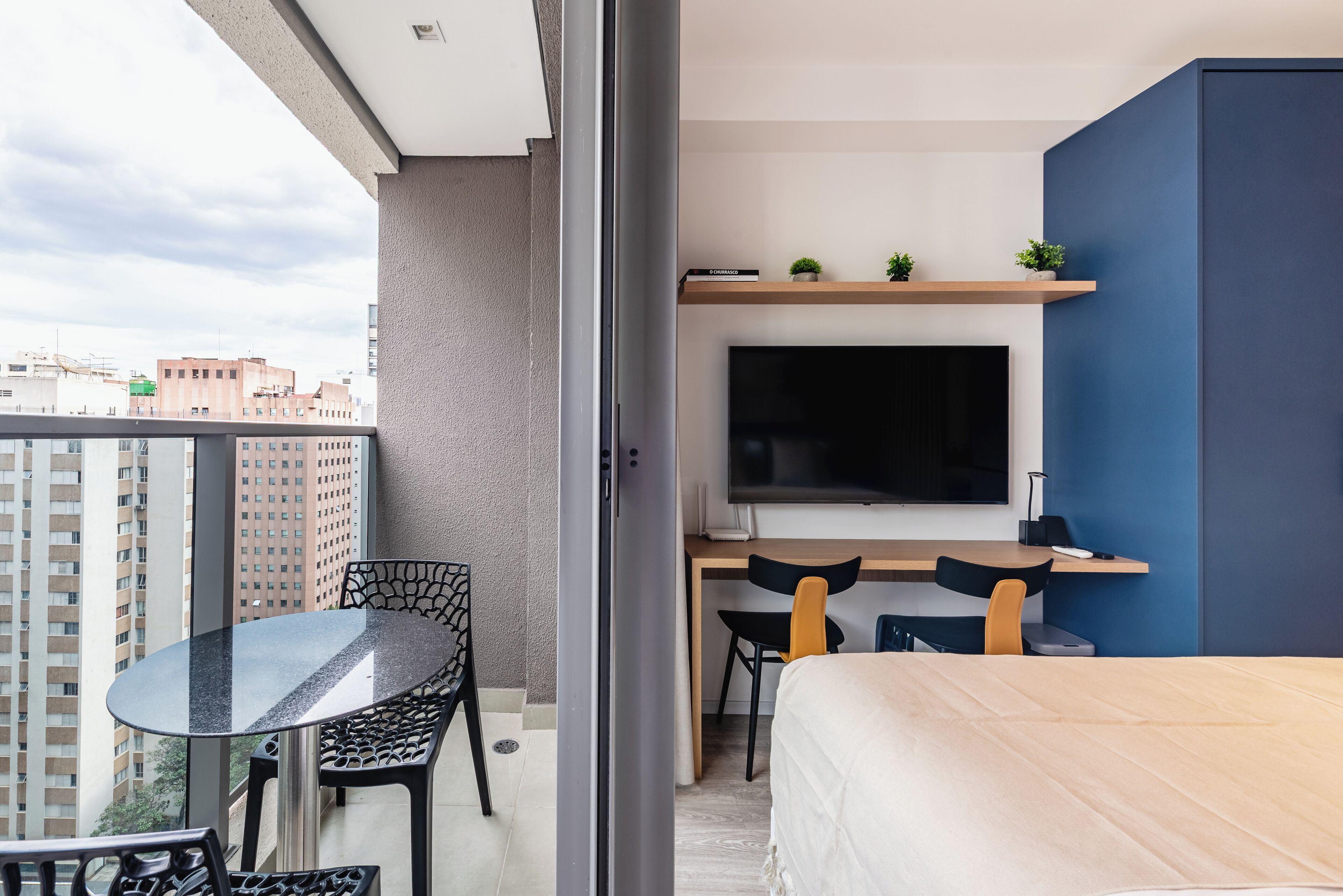 Quarto moderno com cama, escrivaninha com televisão e varanda com mesa e cadeiras.