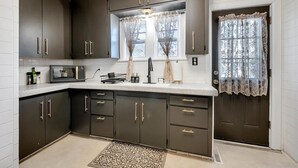 A modern kitchen with dark cabinets, a microwave, and a window with lace curtains. - Clifton Corner: Art Deco charm in College Hill. Walk to the park, dining & shops. 2 king beds. (Wichita)