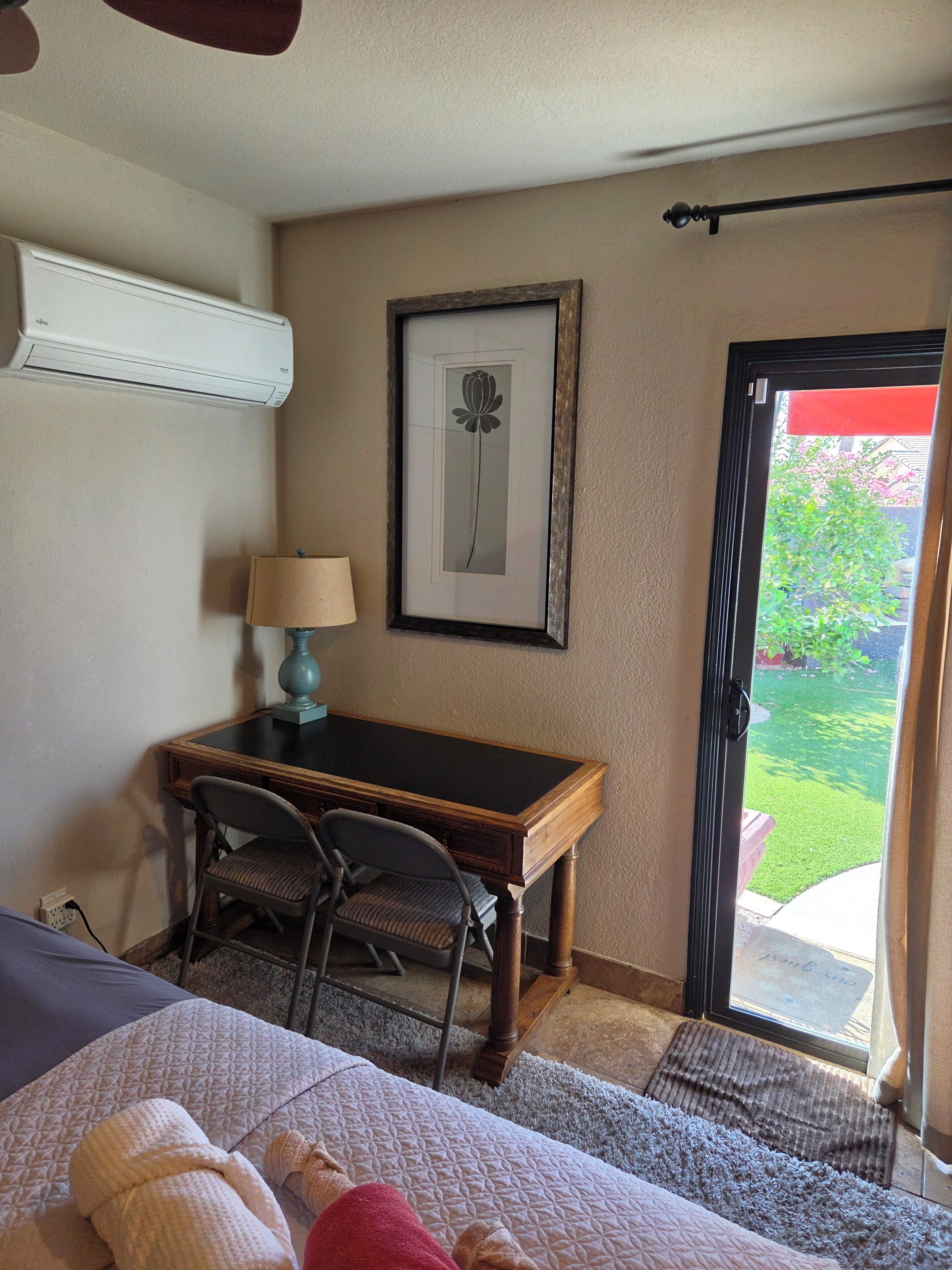 A hotel room with a bed, a desk, a chair, a lamp, a framed picture on the wall, and a door leading to an outdoor area.