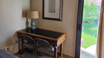 A hotel room with a bed, a desk, a chair, a lamp, a framed picture on the wall, and a door leading to an outdoor area.