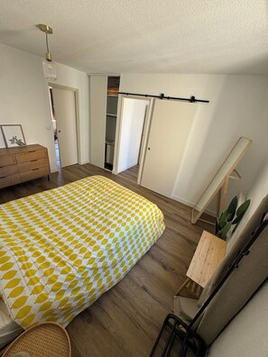 Ein Schlafzimmer mit Bett, Kommode, Schreibtisch, Stuhl, Pflanze und Kleiderschrank.
