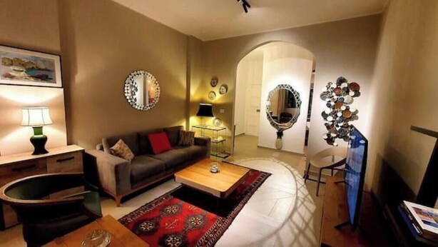 Een woonkamer met een bank, salontafel en een rood vloerkleed. Er staan twee lampen, een spiegel en een kunstwerk aan de muur.
