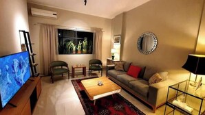 Een woonkamer met een flatscreen televisie, een bank, twee fauteuils, een salontafel en een groot raam met gordijnen.