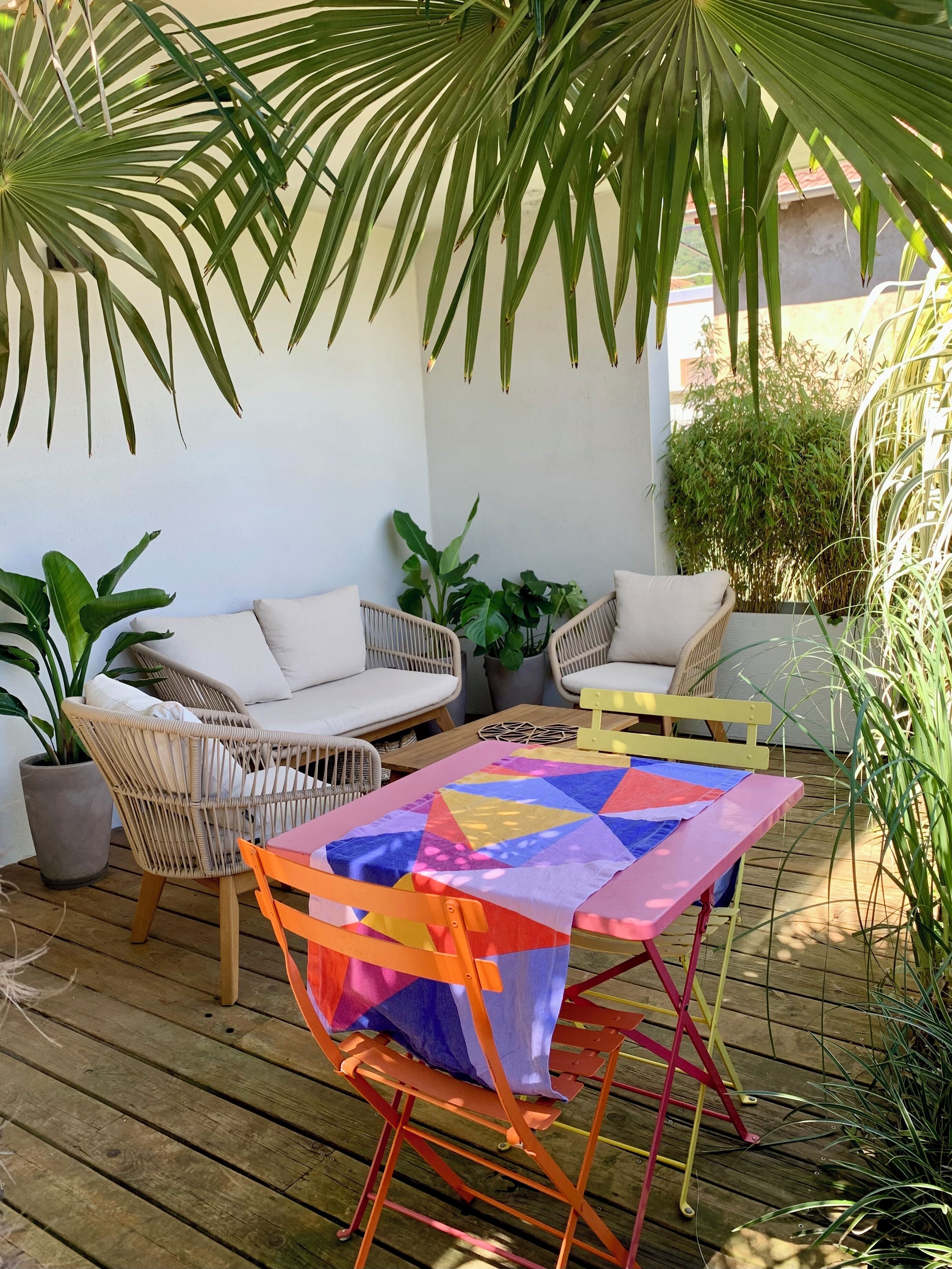 Een terras buiten met rieten meubilair, een kleurrijke quilt op een tafel en diverse potplanten.