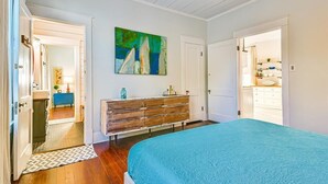 A bedroom with a bed, a dresser, a painting, and a hallway leading to a bathroom.