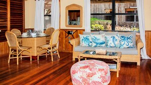 Una habitación con una mesa de comedor lista para cuatro personas, un sofá con cojines decorativos, una mesa de centro de mimbre y un ventanal con vistas a un jardín.