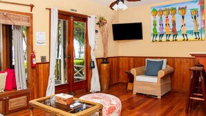 Una habitación con piso de madera, una silla de mimbre, una mesita de centro de vidrio, un televisor montado en la pared y un cuadro que muestra mujeres cargando canastas.