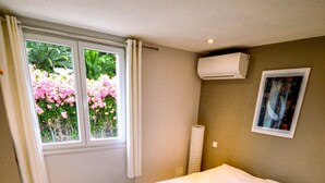 A bedroom with a bed, a window with curtains, an air conditioner, and a framed picture on the wall. - White Cotton (Elne)