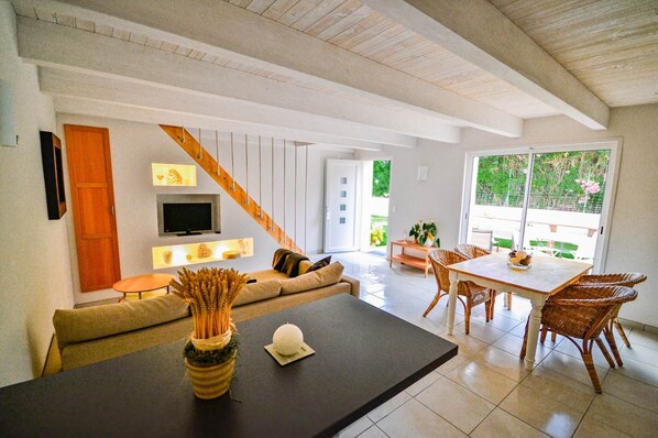 A modern living room with a sofa, a coffee table, a TV, and a dining area with wicker chairs.