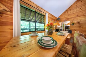 A wooden cabin interior with a dining table set for two, a large window, and a view of greenery outside.