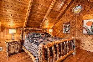 Een slaapkamer in een blokhut met een bed, houten hoofdbord en een raam met uitzicht op bomen.