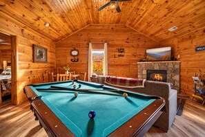 A cozy indoor setting with a pool table, a fireplace, and a view of the outdoors. - Hot Tub with Mountain Views, Fire Pit & Pool Table (Sevierville)