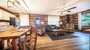 Un salon chaleureux, aménagé dans le style d’un chalet, avec une table à manger en bois, un canapé et des fauteuils. On peut voir une cheminée en pierre et un ventilateur de plafond.