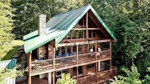 A two-story log cabin with a green roof, multiple balconies, and a stone chimney. - Mountain View Cabin with Hot Tub, Fire Pit & Deck (Sevierville)