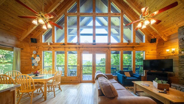A spacious cabin with a high ceiling, wooden walls, and large windows. There is a dining area with a table and chairs, a sofa, and a television. - Mountain View Cabin with Hot Tub, Fire Pit & Deck (Sevierville)