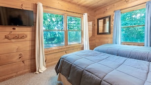 A cabin room with two beds, a TV, and large windows with a forest view. - Mountain View Cabin with Hot Tub, Fire Pit & Deck (Sevierville)