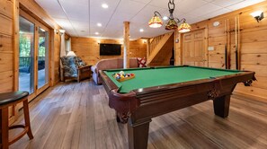 Una mesa de billar en una sala de madera con una escalera, un televisor y un taburete de bar.
