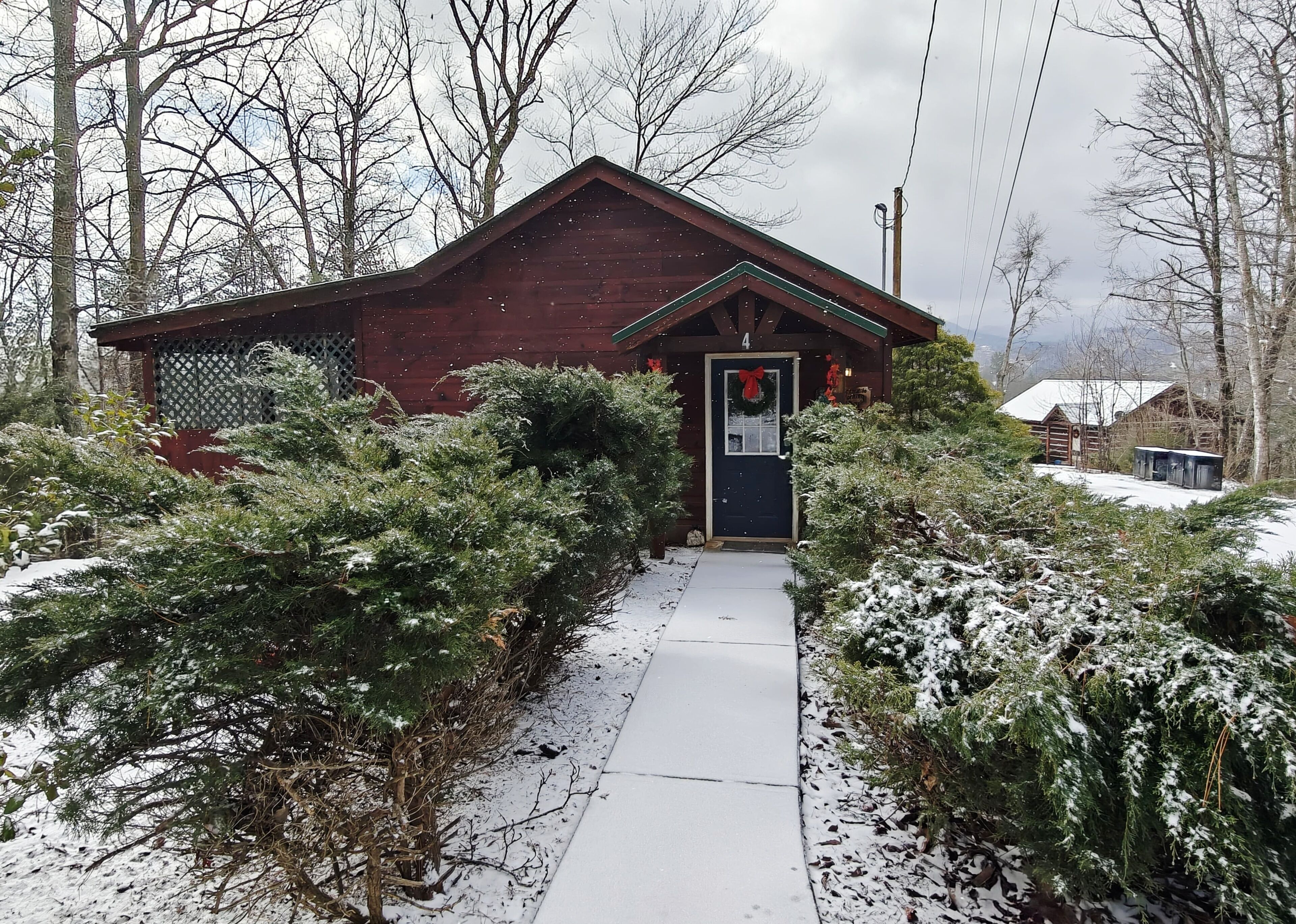 A wooden cabin with a dark door, surrounded by snow-covered bushes and a snow-covered path leading to it.