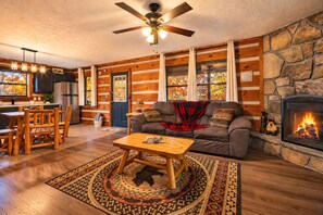 A cozy living room with a fireplace, wooden walls, a dining area with a table and chairs, and a ceiling fan.