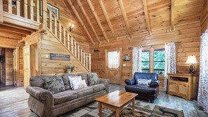 Interior d'una cabana de fusta amb escala, un sofà, una cadira i una taula.