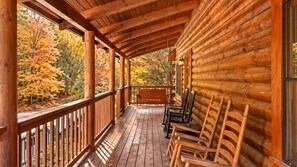 Uma cabana de madeira com varanda, cadeiras de madeira e vista para árvores de outono.
