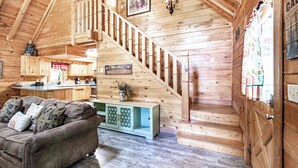 Interior acollidor d'una cabana amb una escala de fusta, un saló amb sofà i coixins, una zona de cuina i una porta amb un cortineta decorativa.