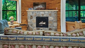A cozy cabin interior with a stone fireplace, a sofa with leaf-patterned cushions, and a view of greenery through large windows.
