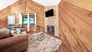 Un acogedor espacio tipo loft con paredes de madera, una chimenea de piedra, un televisor montado en la pared, un sofá y una pequeña mesa con una planta.