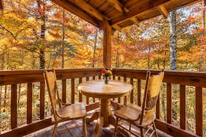 A wooden deck with a round table and two chairs, surrounded by a wooden railing, overlooking a forest with autumn foliage.