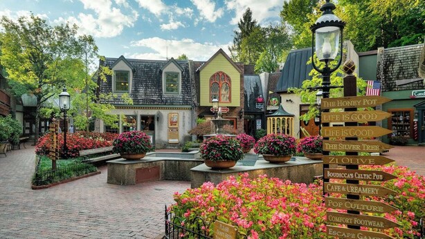 Una pintoresca plaza de pueblo con una fuente central, rodeada de jardines floridos y un cartel que enumera atracciones locales.