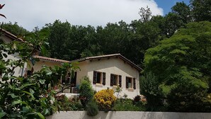 Une maison à deux étages, avec un toit recouvert de tuiles, entourée d’une végétation luxuriante et surplombée d’un ciel bleu clair.