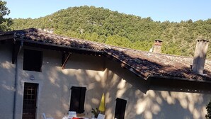 Un bâtiment rustique avec un toit en tuiles, un petit patio avec des chaises en plastique blanches et une pelouse devant.